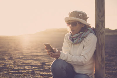 Woman using mobile phone while sitting at beach against sky during sunset