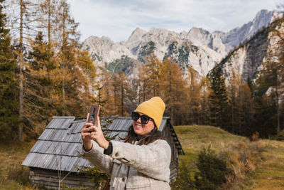 Portrait of happy young woman taking a selfie in front of golden larch trees under mountains