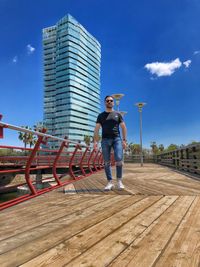 Full length of young man standing on footbridge in city against blue sky