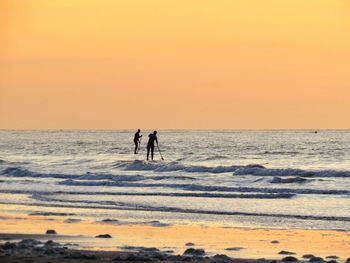 Silhouette of men paddleboarding in sea against sky during sunset