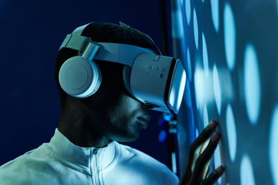 Side view of concentrated young african american in white t shirt and modern vr goggles pointing and touching screen while interacting with virtual object in dark studio with neon illumination