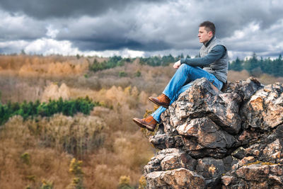 Side view of young man sitting on rock against storm clouds