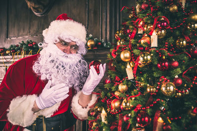 Man wearing santa claus costume holding bauble by christmas tree at home