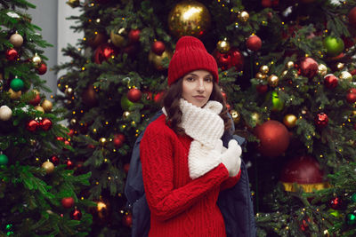 Young woman in a red knitted hat and sweater stands with a gift at the christmas tree