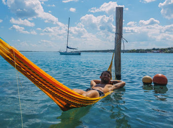 Shirtless young man lying on hammock in sea against cloudy sky during sunny day