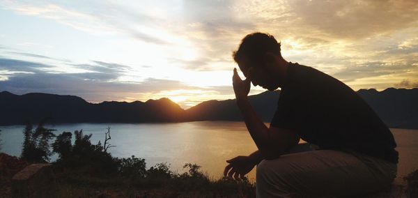 Man sitting by lake against sky during sunset