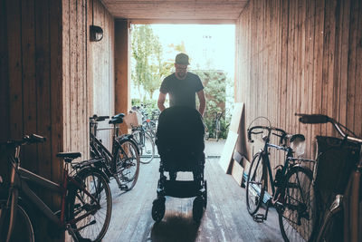 Man pushing baby carriage while walking at corridor amidst bicycles and wooden walls