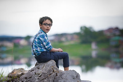 A boy with glasses in a blue plaid shirt sits on a large stone on the shore of a pond