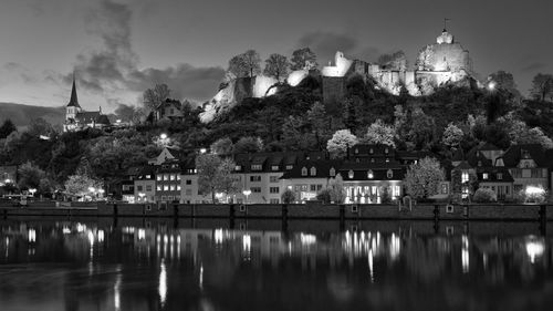 Illuminated buildings by lake against sky in city at dusk