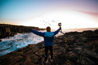 Full length rear view of photographer standing on rock at sea shore against sky during sunset