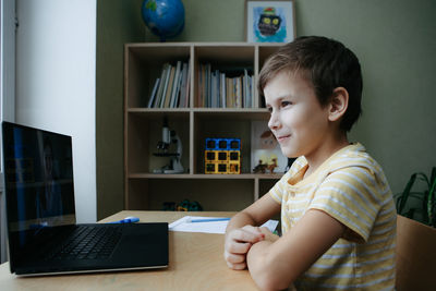 7 years old boy sitting by desk with laptop doing writing task during online lesson. side view