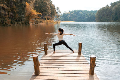 Rear view of woman practicing yoga while standing on pier over lake against trees