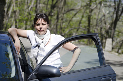 Portrait of confident woman standing by car against trees