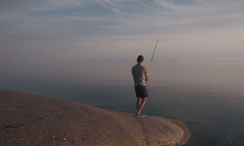 A young man is fishing on the granite shore of a calm sea. back view.