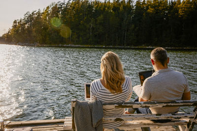 Rear view of couple using laptop while sitting on bench against lake