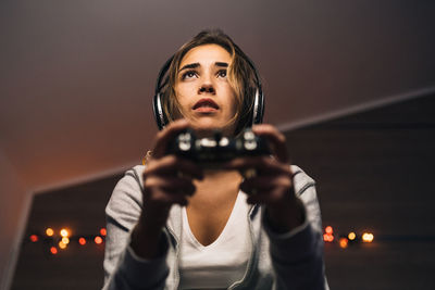 From below concentrated female in headphones and with console sitting in armchair and playing video game while entertaining in evening at home