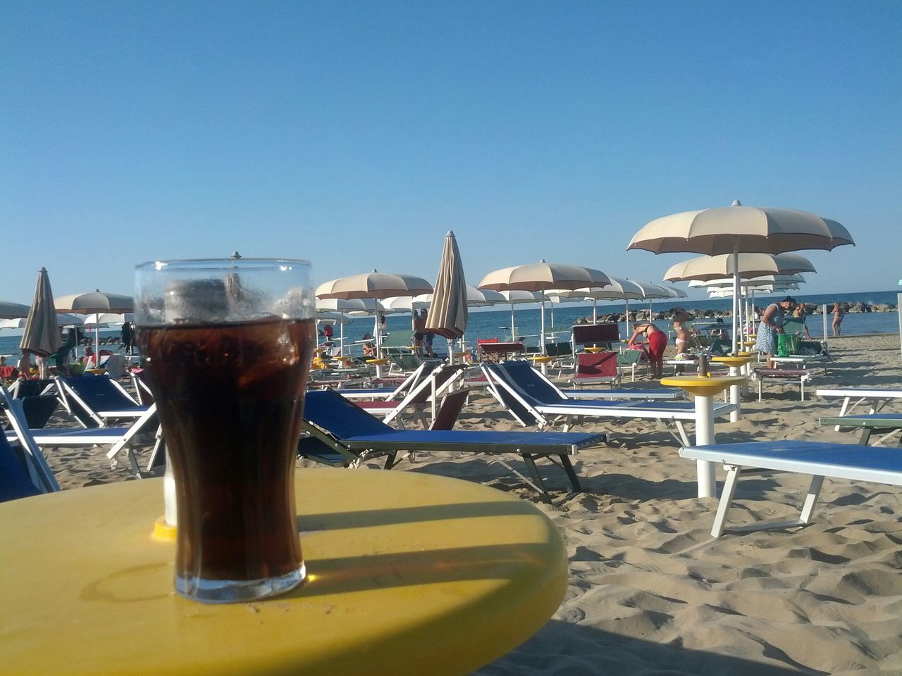 food and drink, drink, table, beach, clear sky, sea, sunlight, refreshment, restaurant, sky, sand, chair, drinking glass, incidental people, shore, outdoors, day, horizon over water, summer, blue