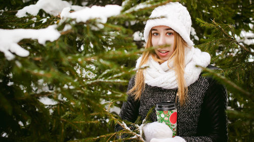 Young woman wearing warm clothing while holding disposable cup amidst trees during winter