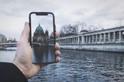 Man holding smart phone over river against sky