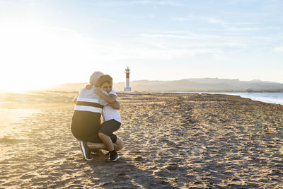 Grandfather and grandson embracing at beach against lighthouse