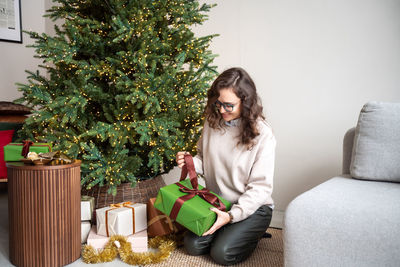 A cute girl with glasses is sitting on the carpet near the christmas tree and unpacking gifts