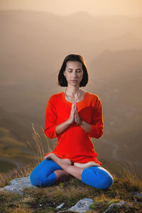 Young caucasian woman meditates in the lotus position in the grass in the mountains near the cliff