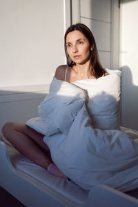 Woman in a white t-shirt is sit on the bed under the blanket in the room im morning