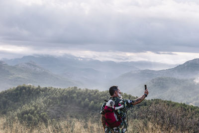 Rear view of man with dog in backpack taking photo high on mountain