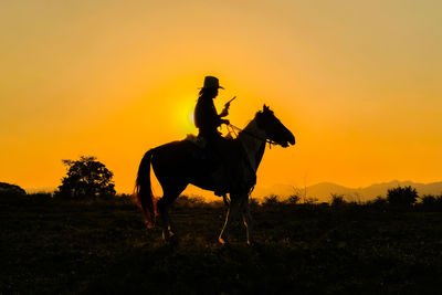 Silhouette man riding horse on field against orange sky