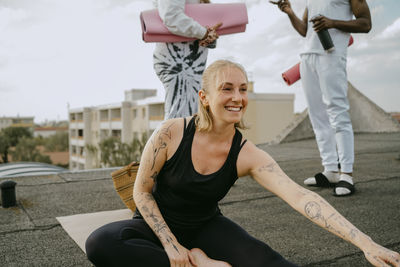 Happy woman practicing yoga while friends talking with each other standing on rooftop