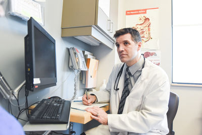 Doctor listening to patient and writing at a desk in a clinic room.