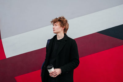 Young man in black coat with cup of coffee in hands looking away outdoors