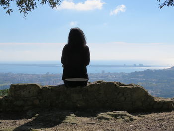 Rear view of woman sitting on retaining wall against sky