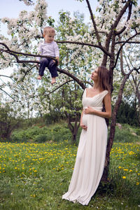 Pregnant woman with long hair and hold her son near the apple trees with white flowers