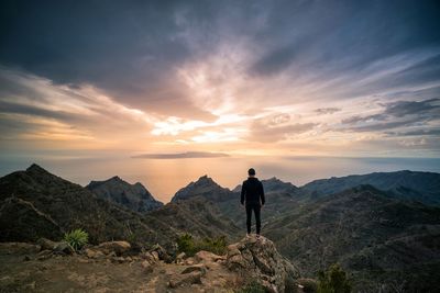 Rear view of mid adult man standing on rock against cloudy sky during sunset