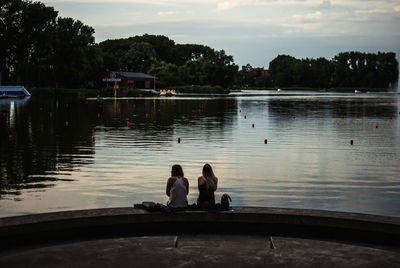 Rear view of women sitting on retaining wall at lakeshore against sky