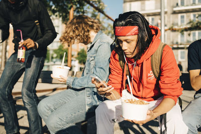 Young man using mobile phone while having take out food by friends in city
