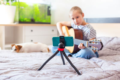 A child plays an guitar at home, looking into a smartphone on a tripod. 