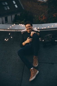 Full length of cheerful man using mobile phone while relaxing on illuminated terrace during party at dusk