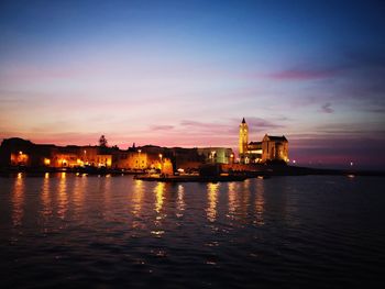 Illuminated buildings by sea against sky during sunset