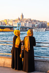 Rear view of men standing by river against buildings in city