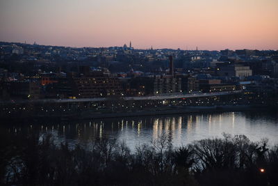 Illuminated city by river against sky at sunset