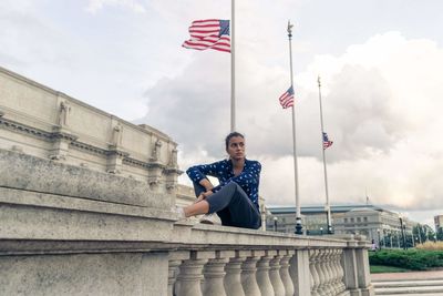 Full length of young woman sitting by american flags on retaining wall