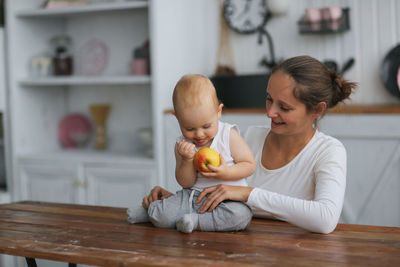 Mother gives the baby an apple, mother hugs her son. real interior, bright kitchen. 