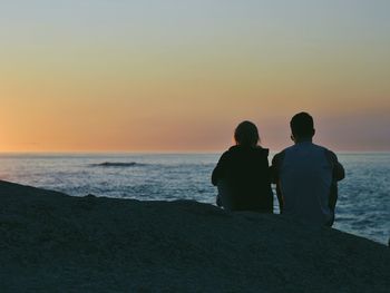 Couple sitting on cliff in front of sea during sunset