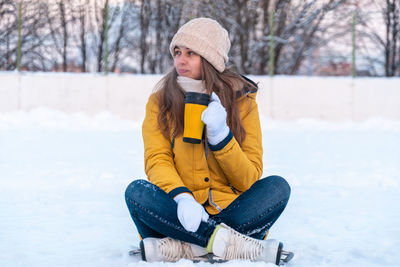 Portrait of young woman in ice skates sitting on the snow, drinking coffee and looking away