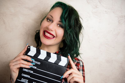 Portrait of smiling young woman holding film slate against wall