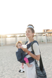 Happy mother feeding milk to daughter at beach against sky during sunset