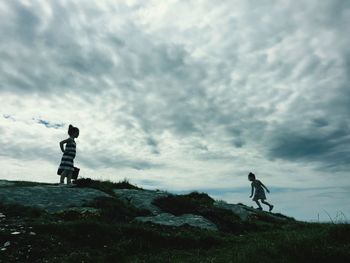 Siblings on rocks at grassy field against cloudy sky