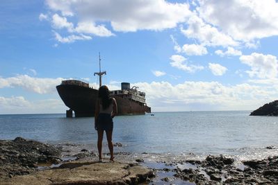 Woman looking at ship while standing on sea shore against cloudy sky